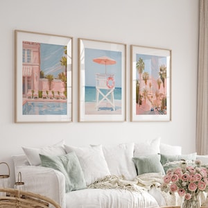 Pode incluir: Três impressões de arte emolduradas retratando cenas costeiras. As impressões apresentam um edifício rosa com piscina, um posto de salva-vidas com guarda-sol e uma rua com palmeiras. A obra de arte tem uma paleta de cores pastel e é exibida acima de um sofá branco.