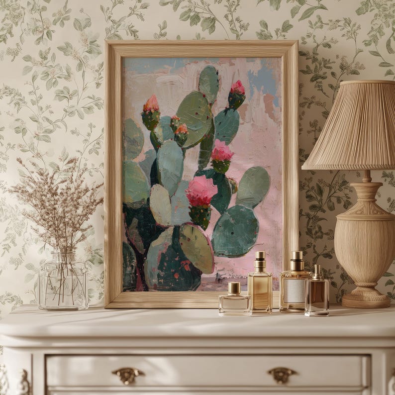 May include: A framed painting of a cactus with pink flowers against a pink and blue background. The painting is on a white dresser with four perfume bottles in front of it.