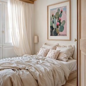 May include: A bedroom with a bed made with a beige linen duvet and pillows. The bed is next to a window with white sheer curtains. A framed print of a cactus with pink flowers hangs on the wall above the bed.