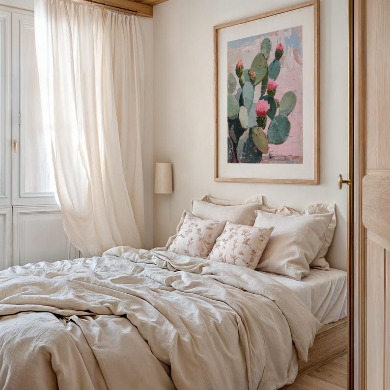 May include: A bedroom with a bed made with a beige linen duvet and pillows. The bed is next to a window with white sheer curtains. A framed print of a cactus with pink flowers hangs on the wall above the bed.