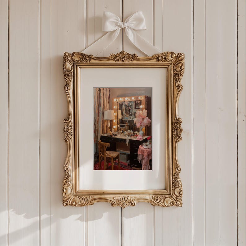 May include: A framed artwork featuring a vanity scene, with a gold ornate frame and a white bow at the top. The artwork depicts a vanity table with a mirror, lights, and various beauty products. The frame is approximately 24 inches tall.