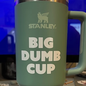 May include: A green stainless steel travel mug with a black handle. The mug has a white logo that says "Stanley" and the text "Big Dumb Cup".