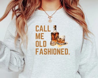 May include: A gray sweatshirt with the text "Call me old fashioned." in brown lettering. The text is accompanied by a graphic of a bottle of whiskey and a glass of whiskey with ice.