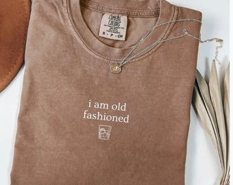 May include: Espresso-colored Comfort Colors t-shirt with the phrase "i am old fashioned" and a cocktail glass graphic. A silver necklace with a pendant rests on the shirt. The shirt is folded and displayed on a white surface.