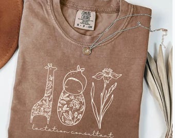 May include: Espresso-colored t-shirt with a white graphic design featuring a giraffe, a baby, a flower, and the text "lactation consultant." The shirt has a crew neck and short sleeves. A silver necklace with a pendant is draped across the shirt.