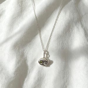 Peut inclure: Un collier en argent composé d'une chaîne fine et d'un petit pendentif en argent de forme organique et irrégulière. Le pendentif présente une surface texturée et martelée, ornée d'une unique petite pierre précieuse vert clair.