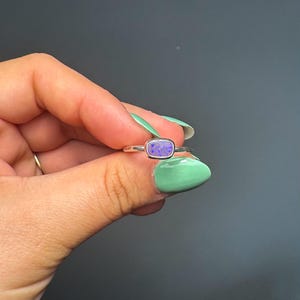 May include: A silver ring featuring a rectangular, purple gemstone with a speckled pattern. The ring is held up against a grey background. This piece of jewellery is a ring.