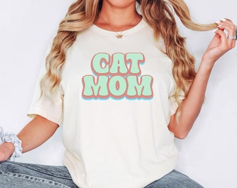 May include: A white t-shirt with the text "CAT MOM" in retro-style green and pink lettering.