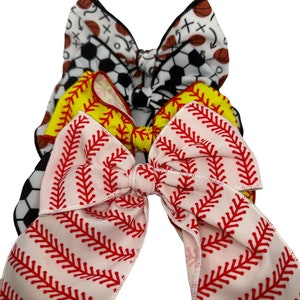 May include: Three hair bows with a sports theme. The top bow is black and white with basketball and soccer ball designs. The middle bow is yellow with baseball stitching. The bottom bow is pink with red baseball stitching.