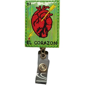 May include: A green, iridescent, embroidered patch with a red heart pierced by a gold arrow. The patch reads "EL CORAZON" and is attached to a silver retractable badge holder.