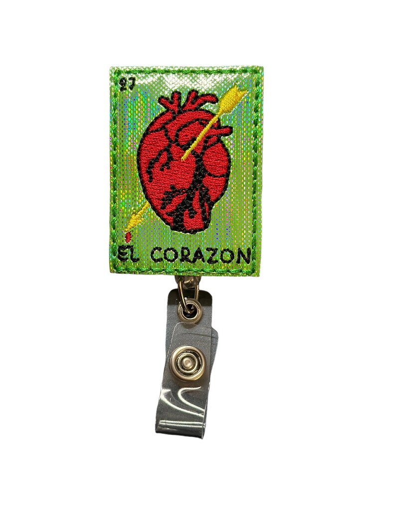 May include: A green, iridescent, embroidered patch with a red heart pierced by a gold arrow. The patch reads "EL CORAZON" and is attached to a silver retractable badge holder.