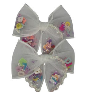 May include: Two white tulle hair bows. Each bow is decorated with colourful alphabet letters and sequins. The edges of the bows are trimmed with a silver, scalloped trim. The bows are tied in the centre with a white ribbon.