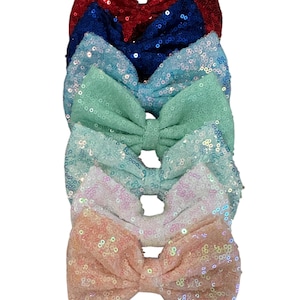 May include: A stack of five sequined hair bows in various colors. The bows include red, blue, light blue, white, and peach. Each bow is tied in a classic bow shape, with a shiny, textured appearance.