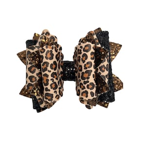 Puede incluir: Un lazo grande para el cabello con un diseño en capas. El lazo presenta un estampado de leopardo beige, detalles negros y dorados con purpurina. El lazo está centrado en un fondo blanco.