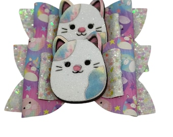 May include: A decorative hair bow featuring two white cat-shaped embellishments with pastel accents. The bow has layers of purple, pink, and blue patterned fabric with glittery accents, creating a whimsical and playful design.