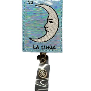 May include: A blue holographic card with a white crescent moon with a face and the text "LA LUNA" and the number "23". The card is attached to a clear plastic retractable badge holder with a gold metal clip.