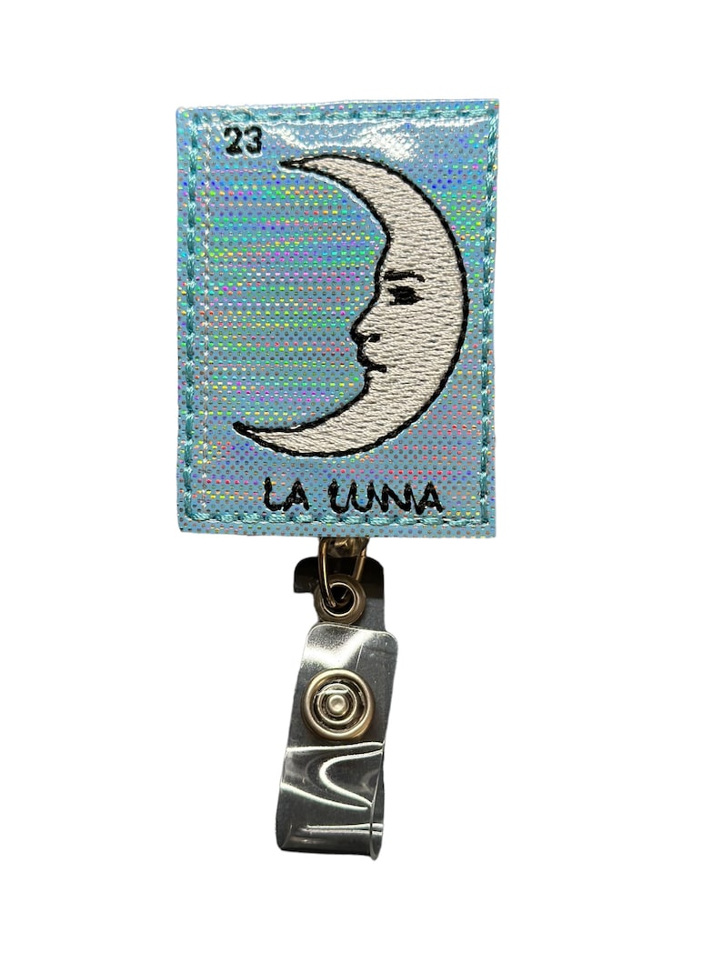 May include: A blue holographic card with a white crescent moon with a face and the text "LA LUNA" and the number "23". The card is attached to a clear plastic retractable badge holder with a gold metal clip.