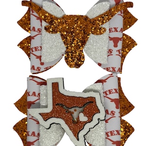 May include: Two hair bows with a white background and orange glitter. One bow features a silhouette of a longhorn steer head. The other bow features a silhouette of the state of Texas with a longhorn steer head inside. Both bows have the words "Texas" and "Longhorns" printed on them.