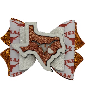 May include: A white and orange bow with a glitter finish. The bow features a cutout of the state of Texas with a longhorn steer inside. The bow is decorated with the word "Texas" repeated on the fabric.