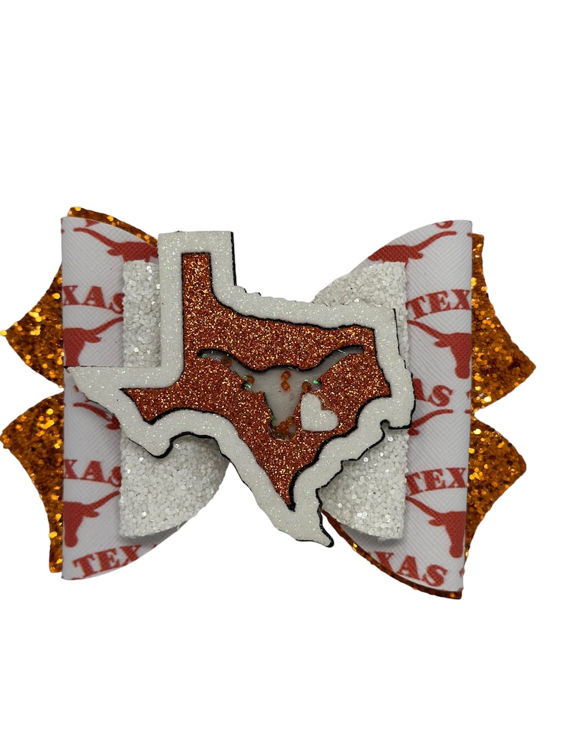 May include: A white and orange bow with a glitter finish. The bow features a cutout of the state of Texas with a longhorn steer inside. The bow is decorated with the word "Texas" repeated on the fabric.