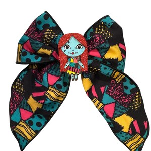 May include: A large, decorative hair bow featuring a patchwork pattern in black, teal, pink, and yellow. The bow is adorned with a glittery Sally character from The Nightmare Before Christmas.