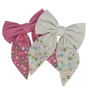 Op de afbeelding: Twee stoffen haarbandjes met een puntige vorm. Een bandje is roze en het andere is wit. Beide bandjes zijn versierd met glimmende pailletten in de vorm van bloemen.