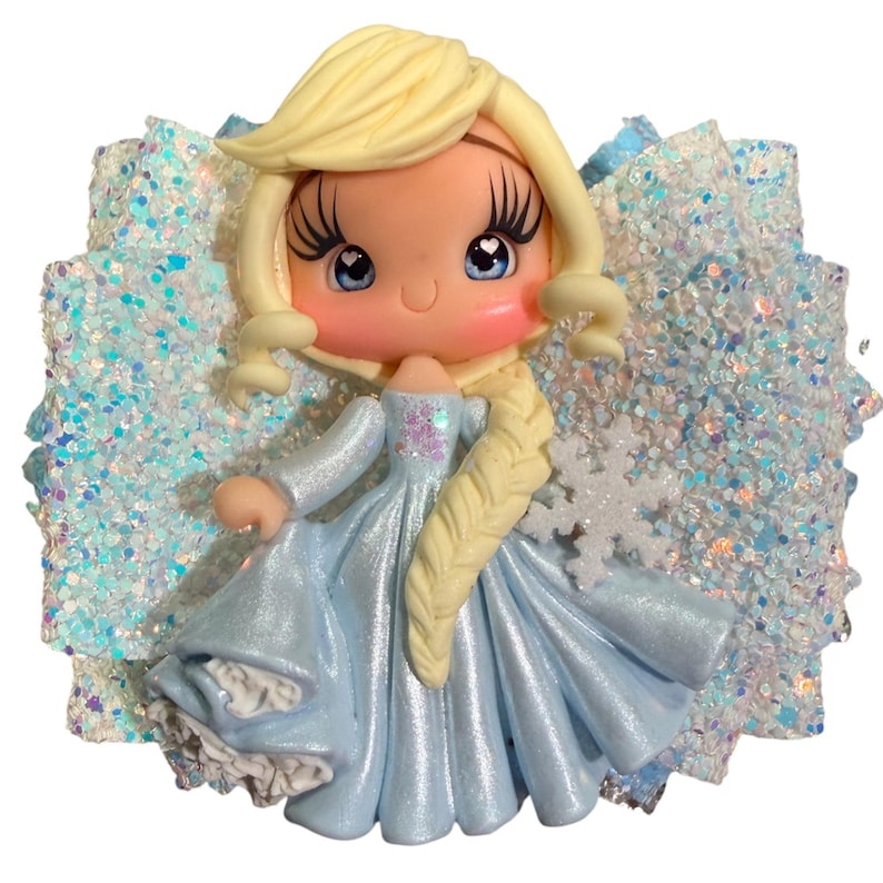May include: A handcrafted hair bow featuring a cartoon character in a light blue dress with a snowflake accent. The character has blonde hair and rosy cheeks, set against a backdrop of glittery blue and white fabric.