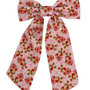 Op de afbeelding: Een roze strik met een Strawberry Shortcake-print. De strik heeft twee lange linten en is in het midden geknoopt.