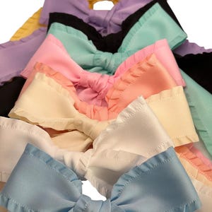 Op de afbeelding: Een stapel van acht handgemaakte stoffen strikken met gekrulde randen in verschillende pastelkleuren, waaronder geel, lavendel, mintgroen, roze, crème, wit en lichtblauw.