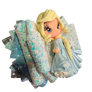 May include: A handmade hair bow featuring a cartoon character with blonde hair and a light blue dress. The bow has layers of glittery light blue and silver fabric, creating a sparkling effect. The character is a stylized representation.