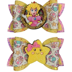 May include: Two glittery hair bows. One features Princess Peach in a gold glitter frame, the other a smiling yellow star with a pink bow. Both bows have a patterned background with cartoon characters and gold and pink accents.