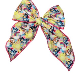 May include: A large, decorative hair bow featuring a repeating pattern of cartoon characters, rainbows, and suns on a yellow background. The bow has a pink trim and is tied in the center.