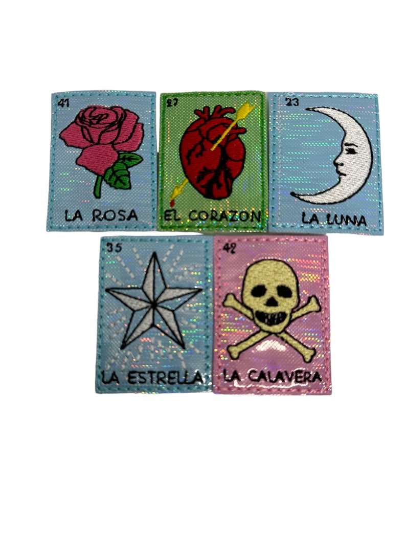 May include: Five colorful embroidered patches with images of a rose, heart with an arrow, crescent moon, star, and skull and crossbones. Each patch has a number and Spanish text, including "La Rosa", "El Corazon", "La Luna", "La Estrella", and "La Calavera".