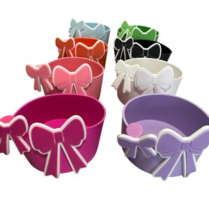 May include: A set of ten colorful plastic containers with a bow design. The containers are in various colors, including pink, purple, white, black, green, orange, and blue.