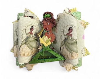 May include: A decorative hair bow featuring a cartoon image of Princess Tiana from Disney's The Princess and the Frog. The bow is green, yellow, and white, with glitter accents. The bow has a layered design with a repeating image of Princess Tiana.