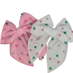 Op de afbeelding: Twee grote haarstrikken, één roze en één witte, worden getoond. Elke strik is versierd met kleine, kleurrijke kralen in roze, groene en witte tinten. De strikken zijn gemaakt van een zacht, fluwelig materiaal.