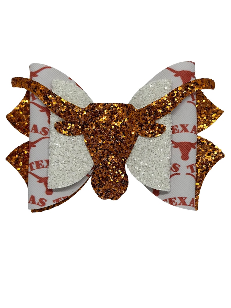 May include: A white and orange glitter bow with a brown glitter Longhorn head in the center. The bow is decorated with the words "Texas" and "Longhorns" in orange and white.