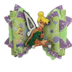 May include: A layered hair bow featuring a cartoon fairy character. The bow has layers of green and purple glitter fabric. The center of the bow features a fairy character with a green dress and yellow hair. The bow is decorated with a floral pattern.