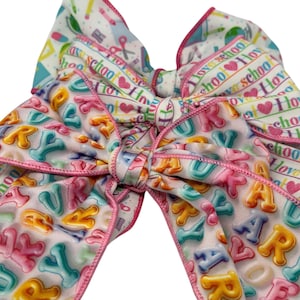 May include: A large, colorful hair bow with a school theme. The bow features a variety of patterns, including the alphabet, pencils, scissors, and the phrase "I love school." The edges are trimmed with pink. A fun accessory for back-to-school.