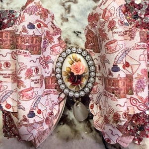 May include: A large, decorative hair bow with a vintage aesthetic. The bow features a cream-colored fabric printed with roses, teacups, and script. A central oval brooch with a floral design and pearl accents adds elegance. The bow's edges are trimmed with glitter.