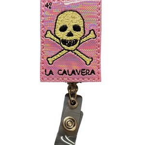May include: Pink fabric badge holder with a gold skull and crossbones design and the text "LA CALAVERA". The badge holder has a clear plastic retractable clip.