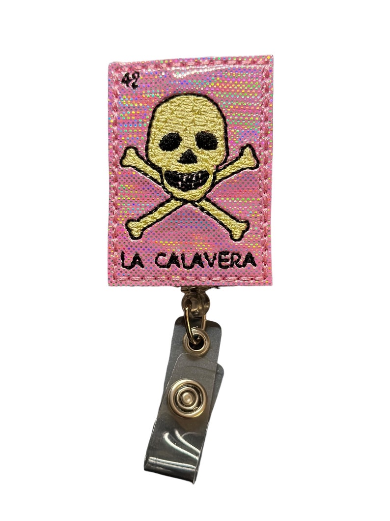 May include: Pink fabric badge holder with a gold skull and crossbones design and the text "LA CALAVERA". The badge holder has a clear plastic retractable clip.