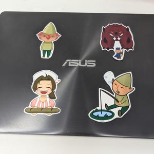 May include: Four colorful stickers on a dark gray laptop. The stickers feature cartoon characters: a gnome, a baker, a monster, and a fisherman. The gnome wears a green hat and outfit. The baker holds bread. The fisherman is fishing.