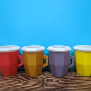Pu&ograve; includere: Quattro tazze geometriche colorate, rosse, oro, viola e gialle, ciascuna con bordo e manico bianchi. Le tazze sono disposte su una superficie di legno contro uno sfondo blu brillante.