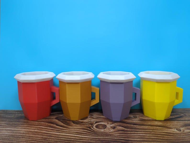 Pu&ograve; includere: Quattro tazze geometriche colorate, rosse, oro, viola e gialle, ciascuna con bordo e manico bianchi. Le tazze sono disposte su una superficie di legno contro uno sfondo blu brillante.