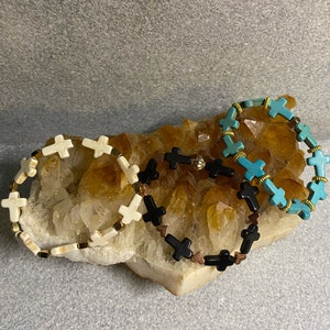 May include: Three bracelets with cross charms. One bracelet has white cross charms, one has black cross charms, and one has turquoise blue cross charms.