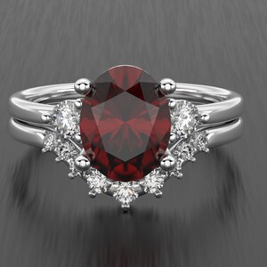 Peut inclure: Ensemble de bagues en argent avec une grosse pierre rouge foncé de coupe ovale au centre. La bague est flanquée de plus petites pierres précieuses claires et repose sur un anneau assorti. Design élégant pour occasions spéciales.