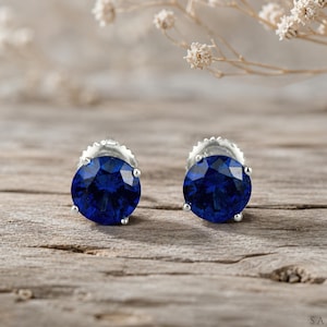 May include: A pair of round-cut, deep blue stud earrings set in silver-toned metal prongs. The earrings feature faceted stones that reflect light, showcasing a vibrant sapphire-like colour. Each stud is secured with a matching silver-toned metal backing.
