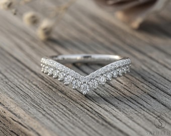 May include: A silver-colored ring with a V-shaped design. The ring is encrusted with numerous small, clear gemstones. The ring is set against a wooden surface, with a blurred background.