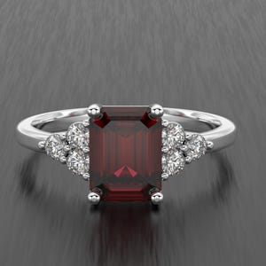 May include: A silver ring featuring a rectangular, deep red gemstone as the centerpiece. The ring is accented with smaller, round, clear gemstones on either side of the main stone, creating a sparkling, elegant design. The ring is set against a dark background.